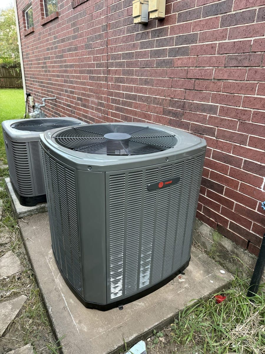 New Trane outdoor condenser on a concrete pad beside a brick house, with a second outdoor unit behind it